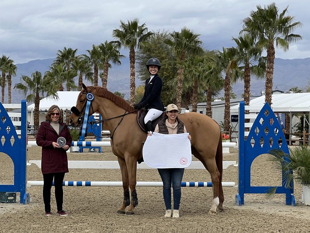 Horse show winner on tan horse with blue jumps, rider and two people holding award ribbon outdoors