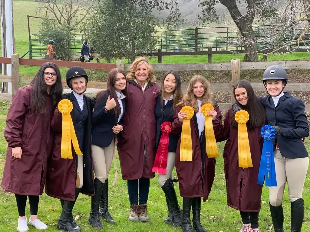 Seven people in maroon robes and riding helmets pose outdoors, holding colorful ribbons and rosettes.