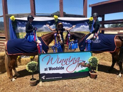 Winners on horses holding ribbons beside a “Wining at Woodside” sign at an outdoor arena