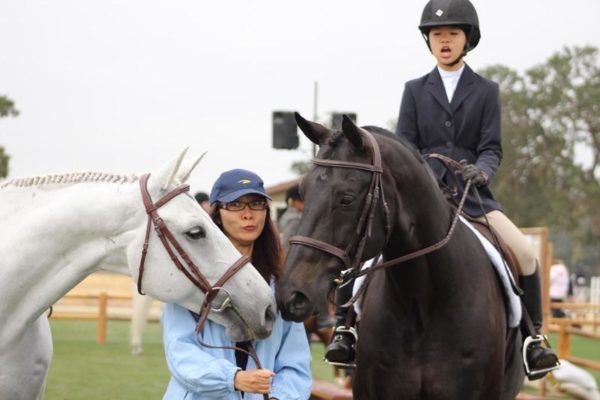 Two horseback riders in helmets on a white and black horse, meeting nose to nose outdoors.