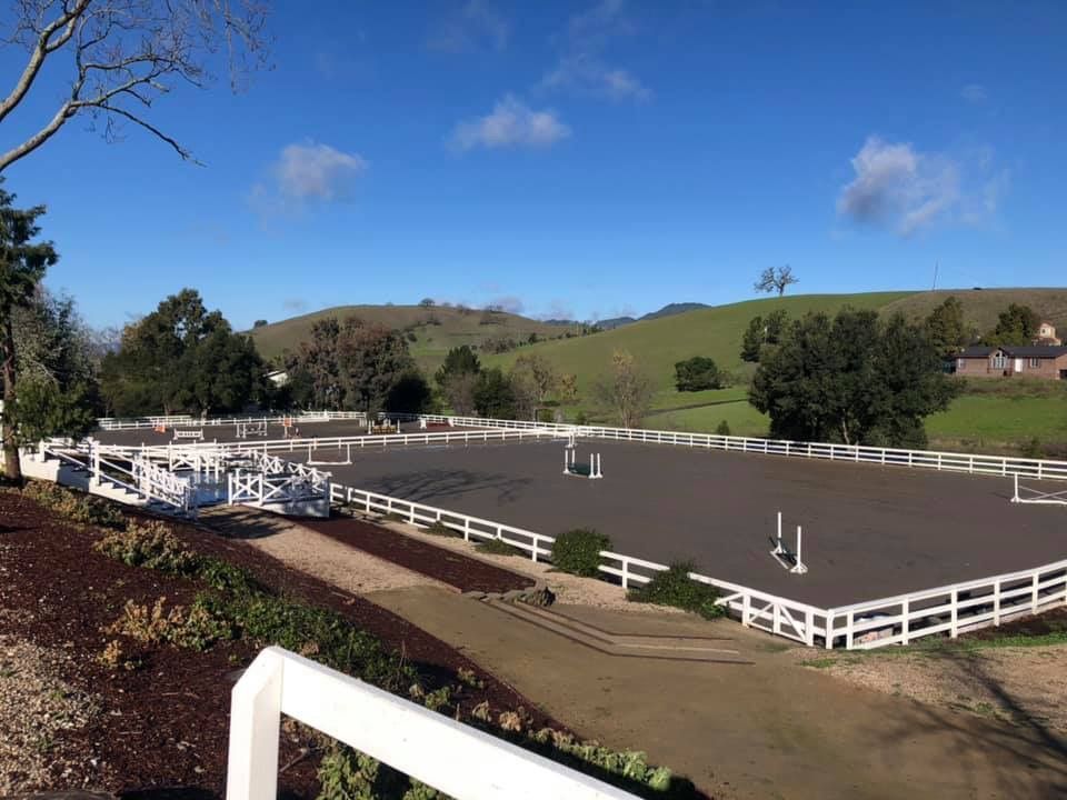 White-fenced outdoor riding arena with jumps under a blue sky, surrounded by grassy hills.