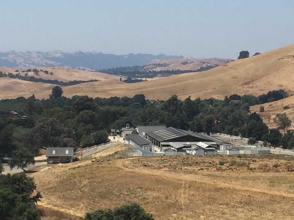 Rural valley with golden hills, trees, and a small farm complex under a hazy sky