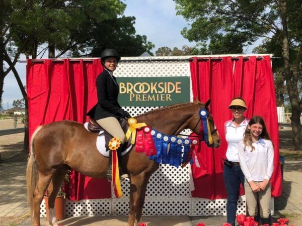 Person on horse posing with two people in front of a Brookside Premier backdrop and red drapes