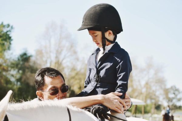 Child in a black riding helmet sits on a white horse while an adult stands beside them outdoors.