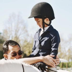 Person in a black helmet sits on a white horse while another person helps adjust the reins outdoors.