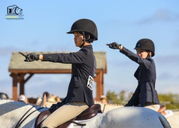 Two riders in black helmets point ahead on white horses during an equestrian event.