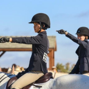 Two riders in black helmets point forward on white horses during an equestrian parade