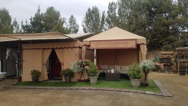 Tan outdoor tented seating area with potted plants and stacked pallets in a garden setting.