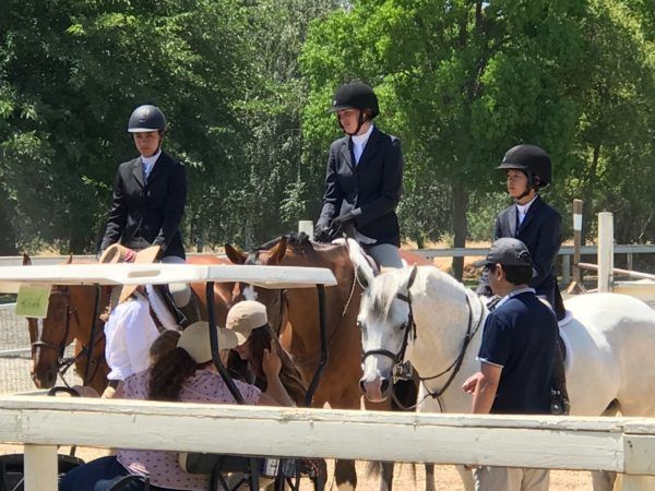 Riders on horses in helmets wait by a fence at an outdoor equestrian arena.