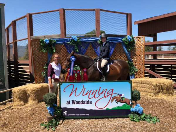 Person with horse and rider beside “Winning at Woodside” sign and hay bales at an outdoor event