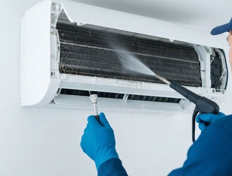 Person cleaning a wall-mounted air conditioner with a spray bottle and brush, wearing blue gloves