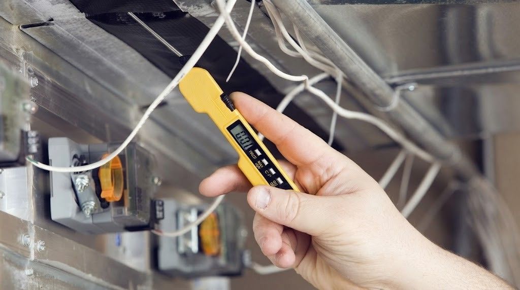Hand holding a yellow utility knife near exposed electrical wiring and metal ceiling panels