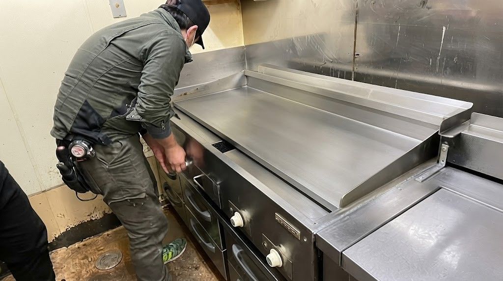 Worker cleaning a stainless steel kitchen grill with open lid in a commercial kitchen