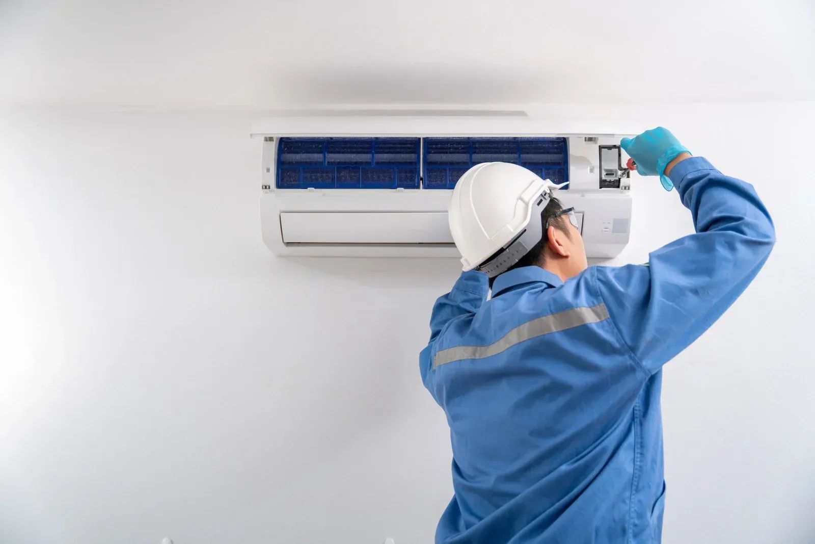 Technician in blue uniform inspecting a wall-mounted air conditioner with a tool