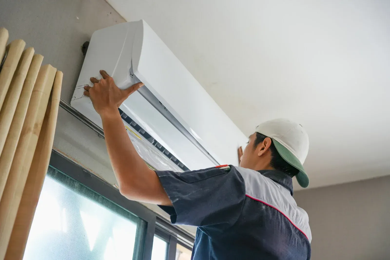 Technician installing a wall-mounted air conditioner near a window.