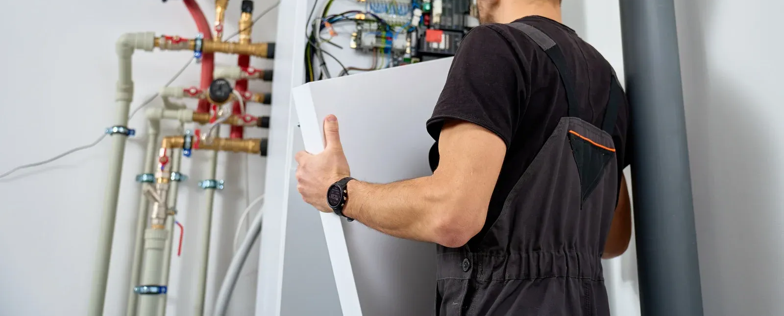 Technician carrying a white panel beside exposed pipes and a large gray tank in a utility room