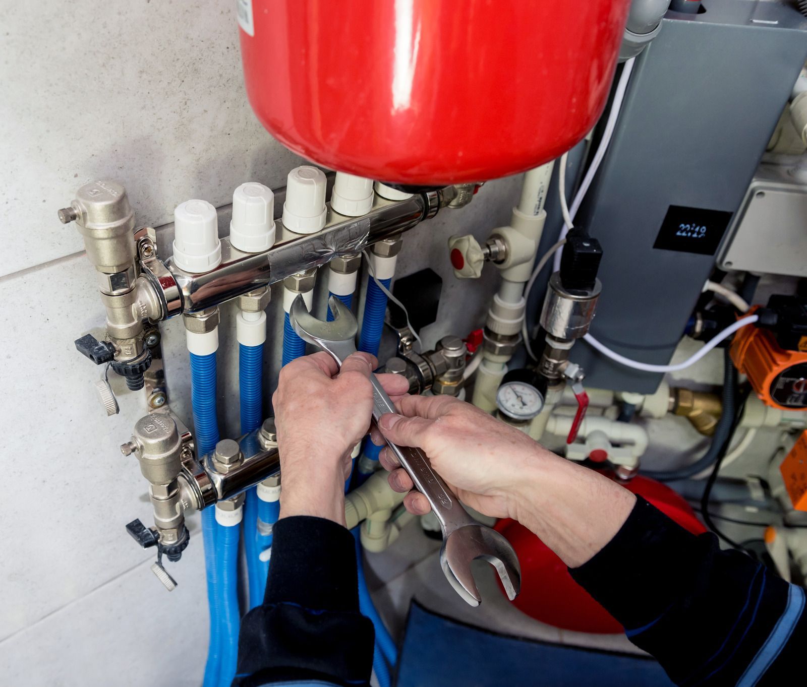 Hands working on blue and white pipes beneath a red tank in a mechanical room