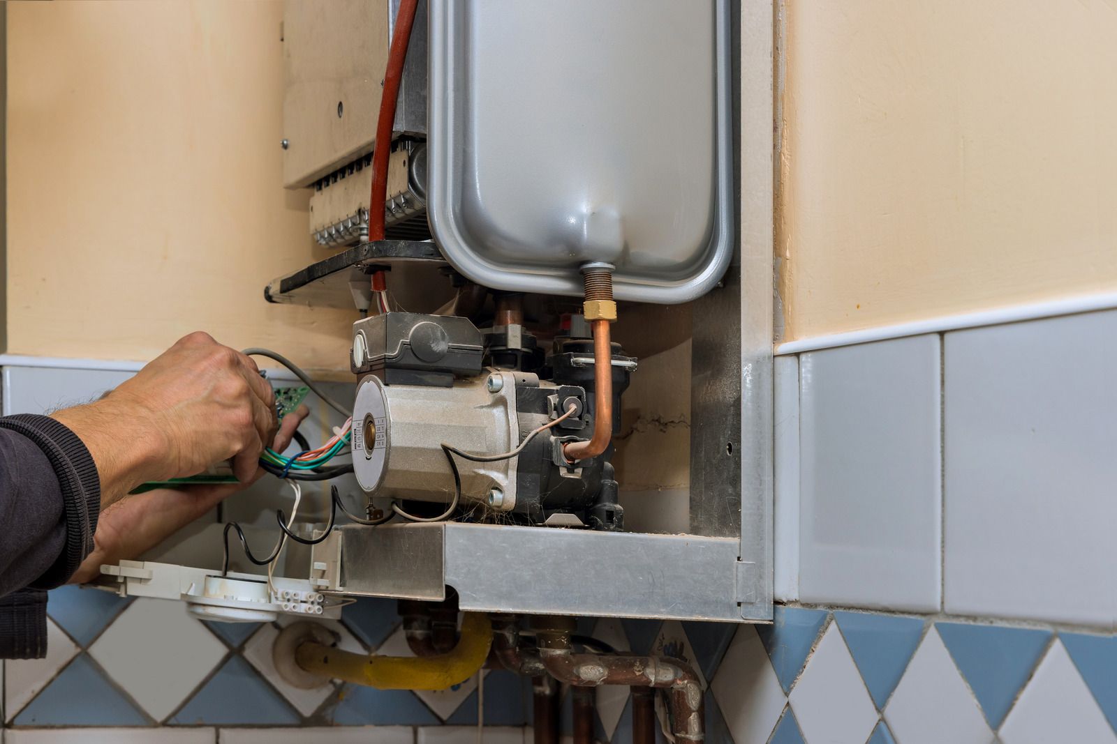 Hand adjusting a wall-mounted gas water heater with pipes and tank in a tiled room
