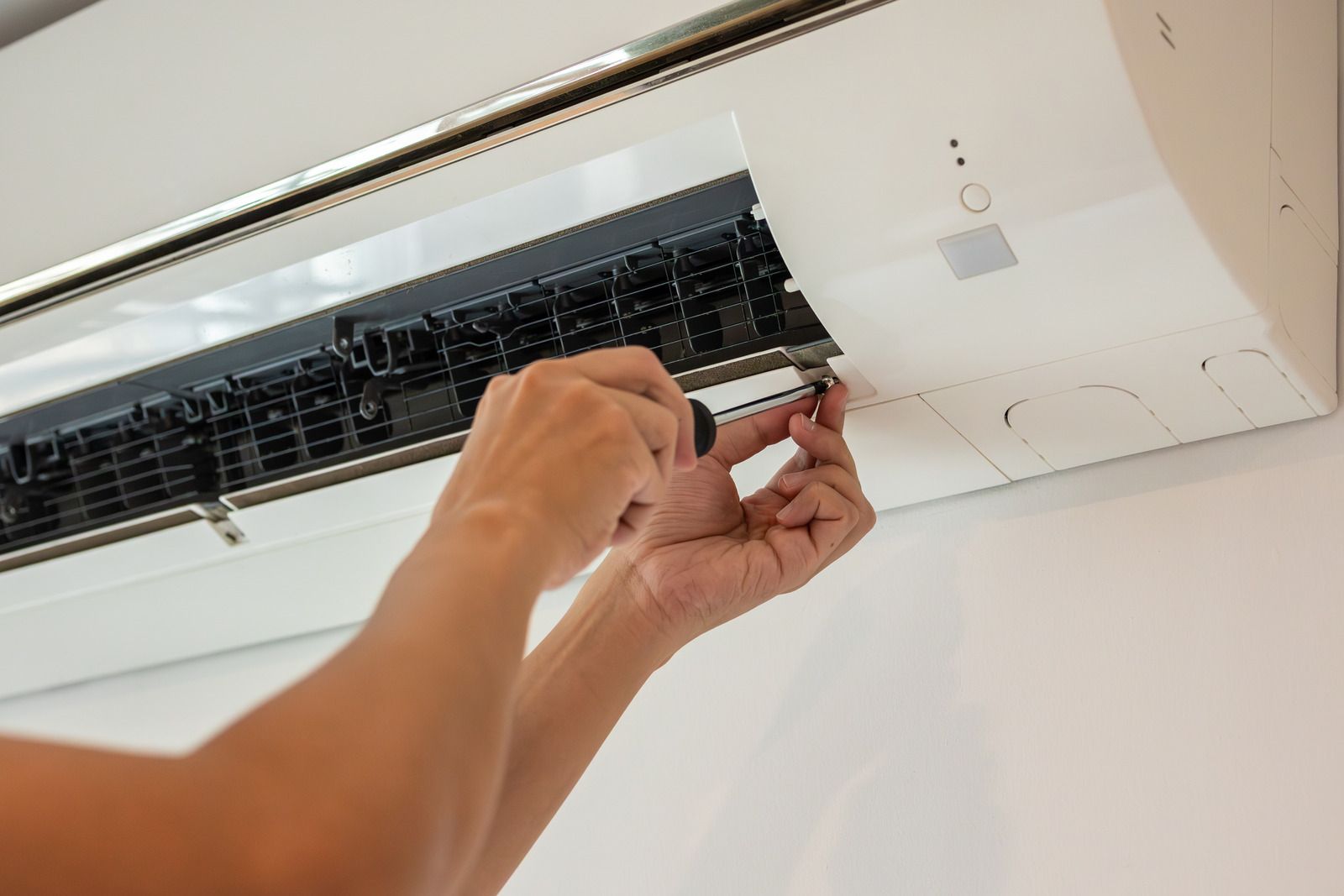 Hands adjusting a white wall-mounted air conditioner vent with a screwdriver