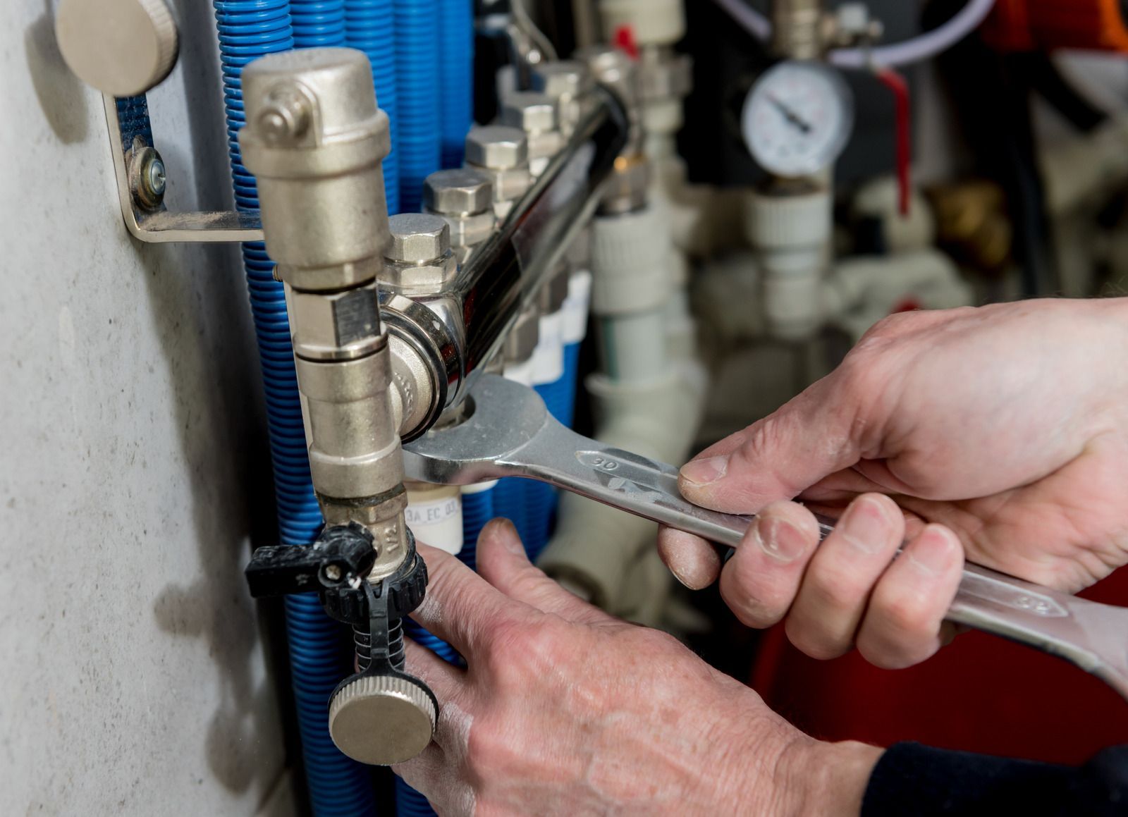 Hands using a wrench to tighten plumbing valves near blue pipes and a pressure gauge.
