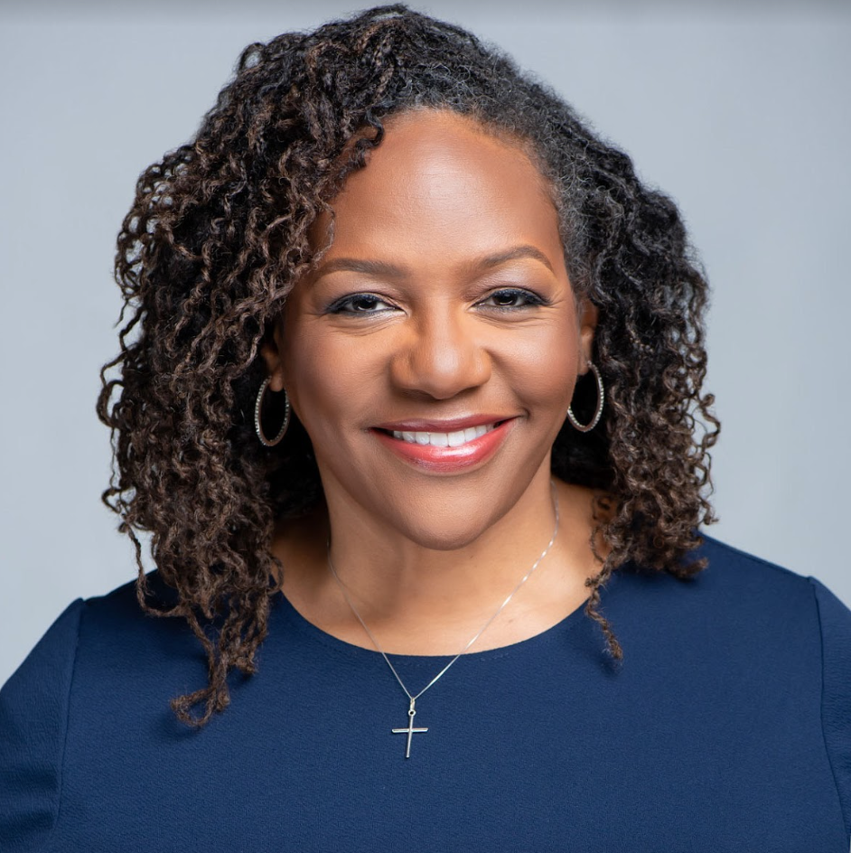 A woman wearing a blue shirt and a cross necklace smiles for the camera.