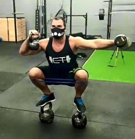 A man wearing a mask is squatting on a kettlebell in a gym.