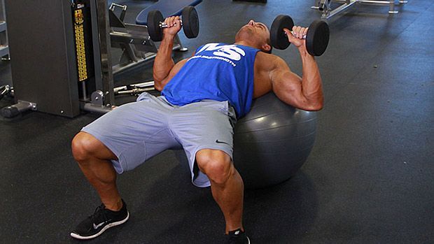A man is sitting on a pilates ball while lifting dumbbells in a gym.