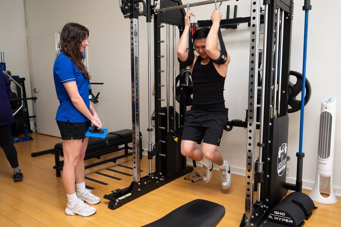 Person doing pull-ups on a cable machine while a trainer watches in a gym.