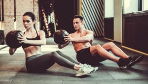 A man and a woman are doing exercises with medicine balls in a gym.