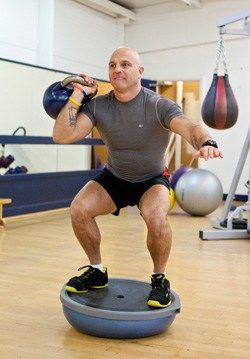 A man is squatting on a balance ball in a gym.