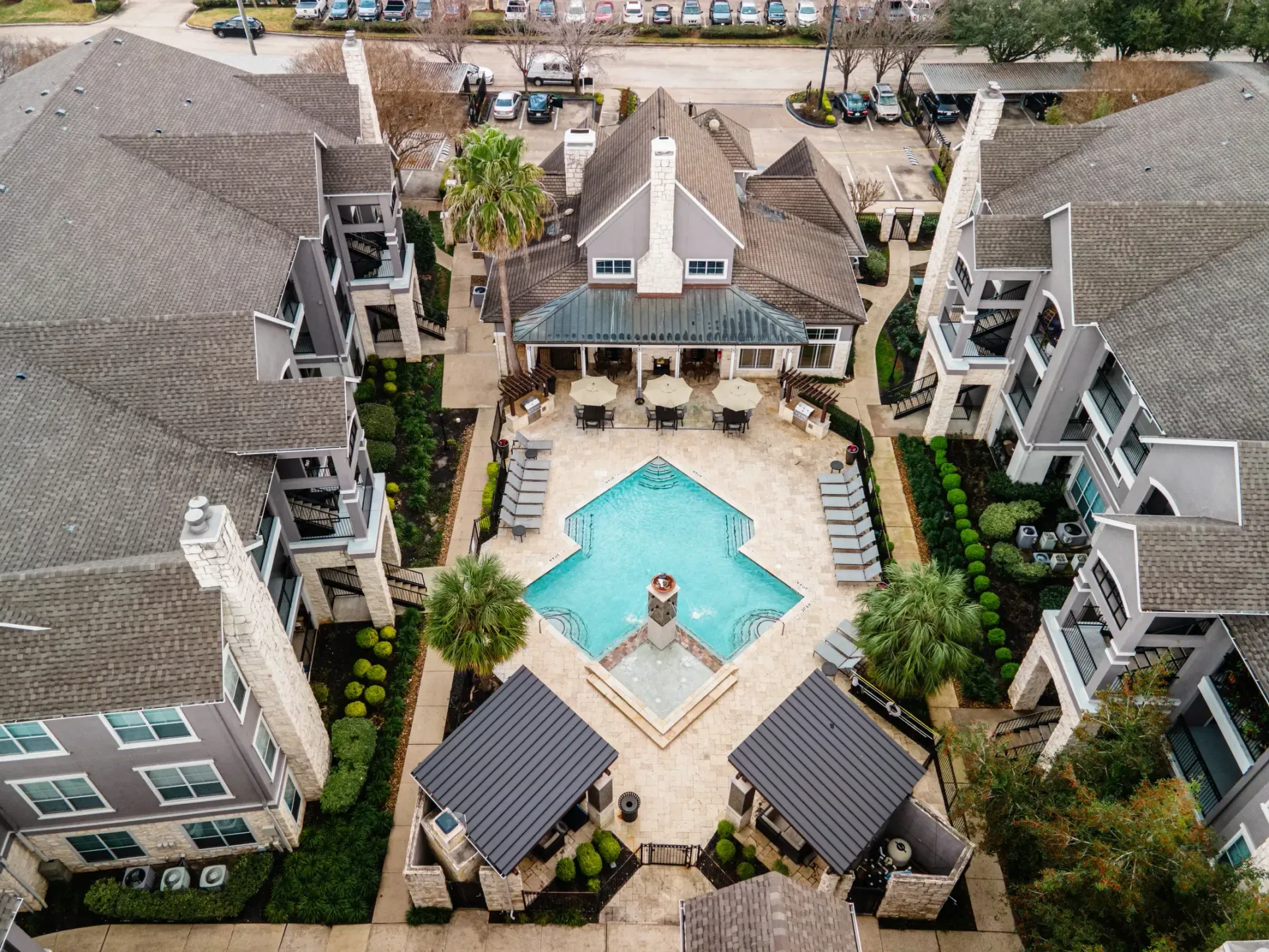 Aerial view of an apartment complex with a pool, central clubhouse, and surrounding buildings with gray roofs and tan walls.