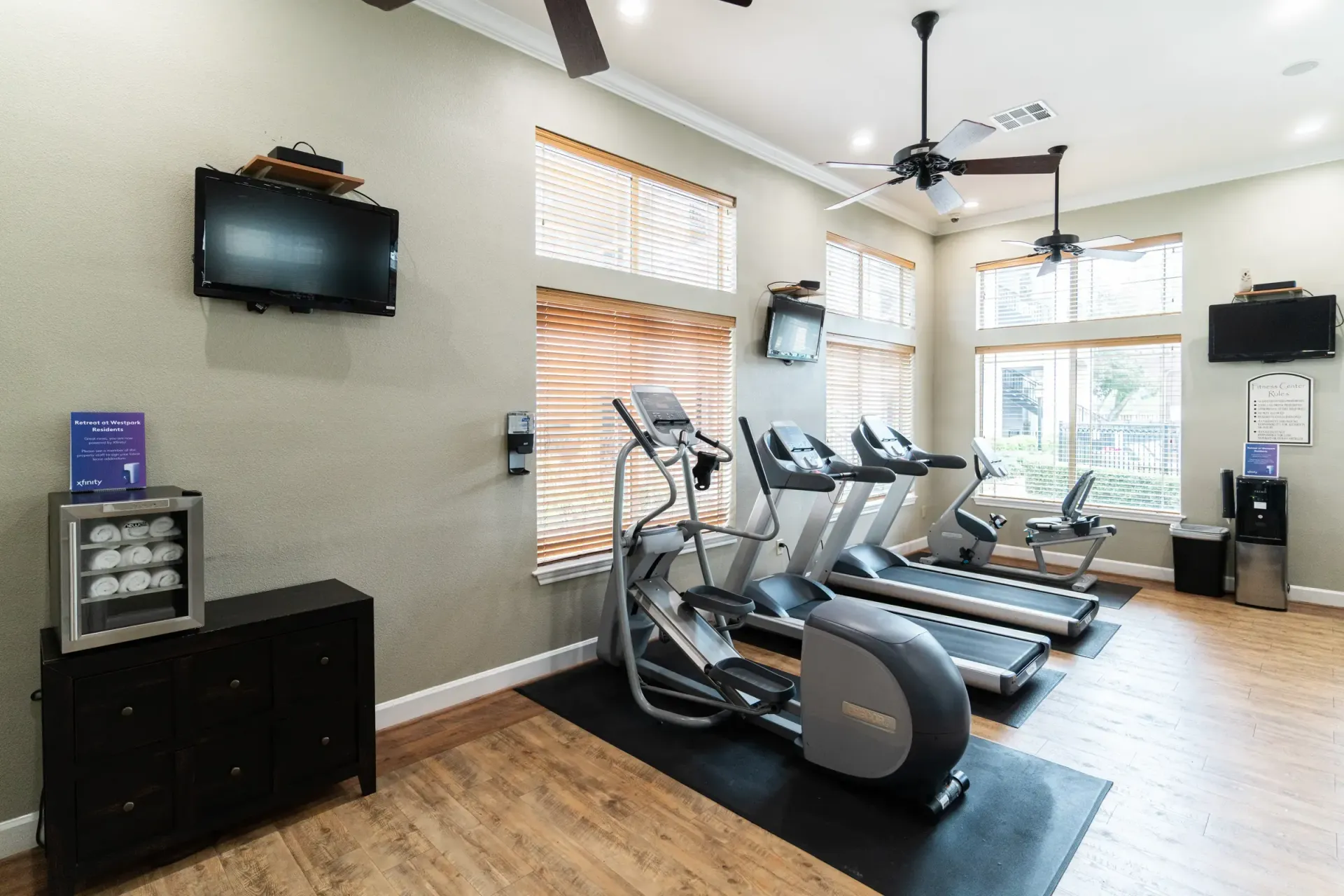 Gym with treadmills, elliptical, and TVs. Windows with shades, ceiling fans, and a storage cabinet.