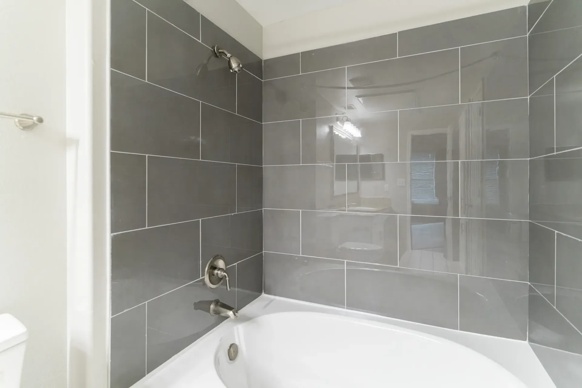 Gray tiled shower with white bathtub and silver fixtures.