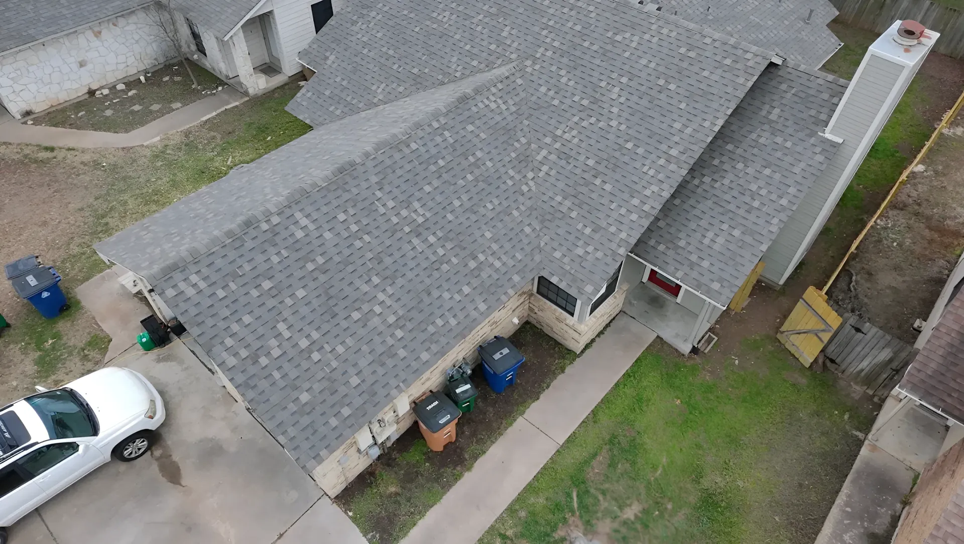 Aerial view of a gray-roofed house with a driveway, parked white car, trash bins, and small yard.