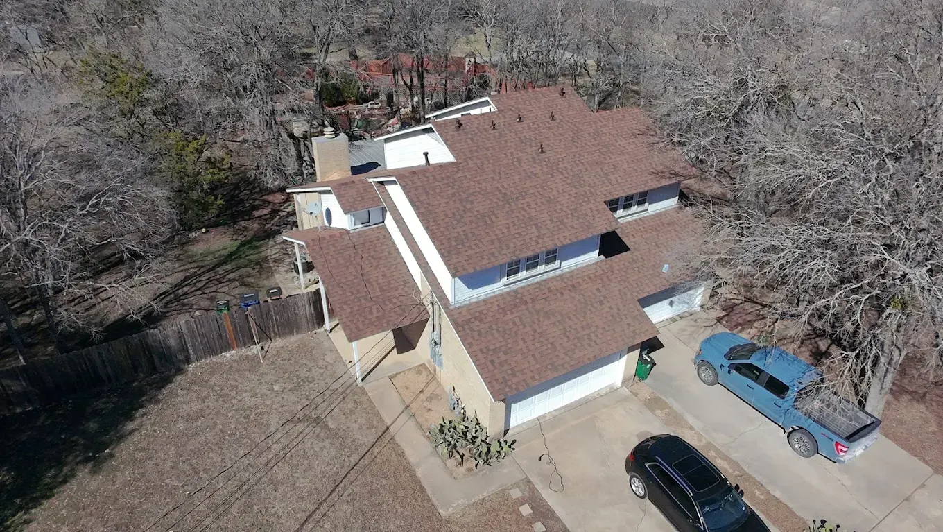 Aerial view of a tan house with a brown roof, surrounded by trees and parked cars in a driveway.