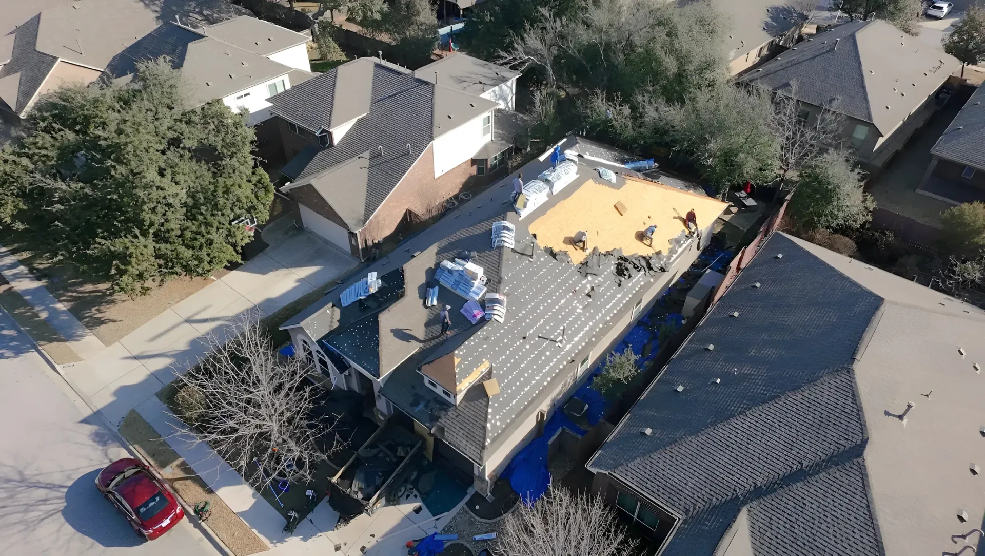 Aerial view of a house roof being repaired, with workers and materials on the rooftop in a suburban neighborhood.