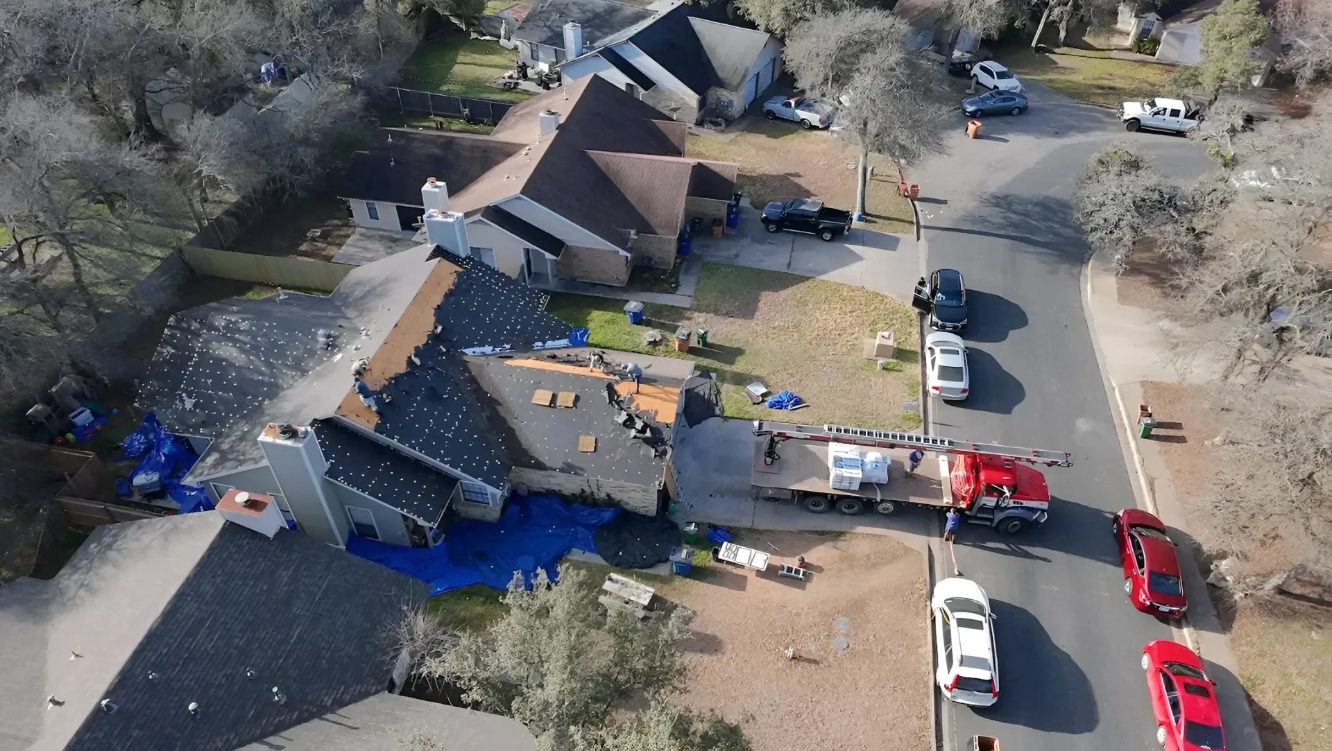 Aerial view of a house with severe roof damage, debris scattered around, and emergency vehicles parked nearby.