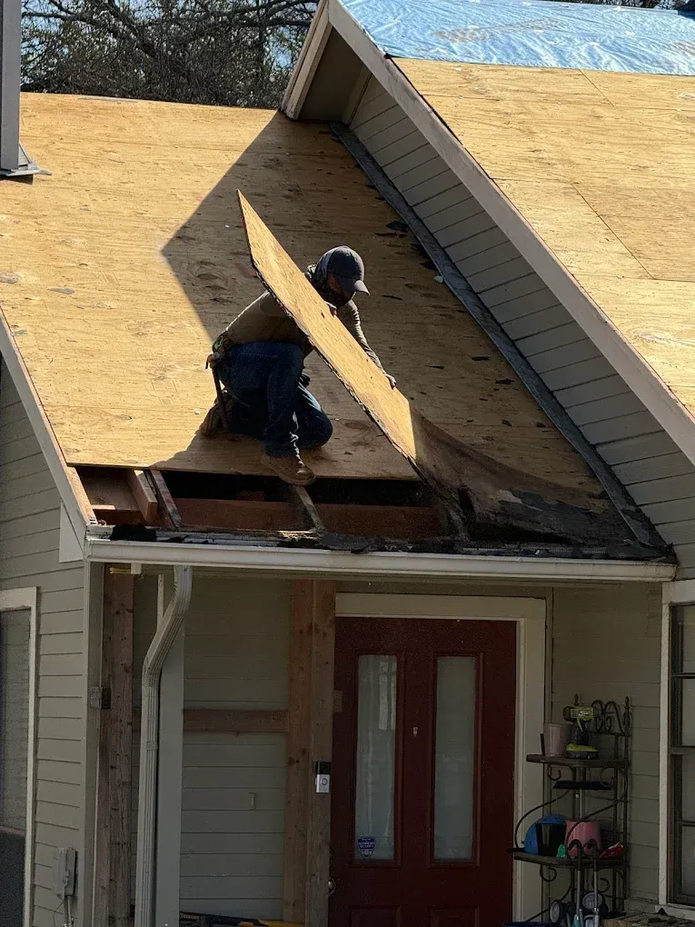 Two workers removing roof shingles on a house porch roof, with plywood exposed above the front door.