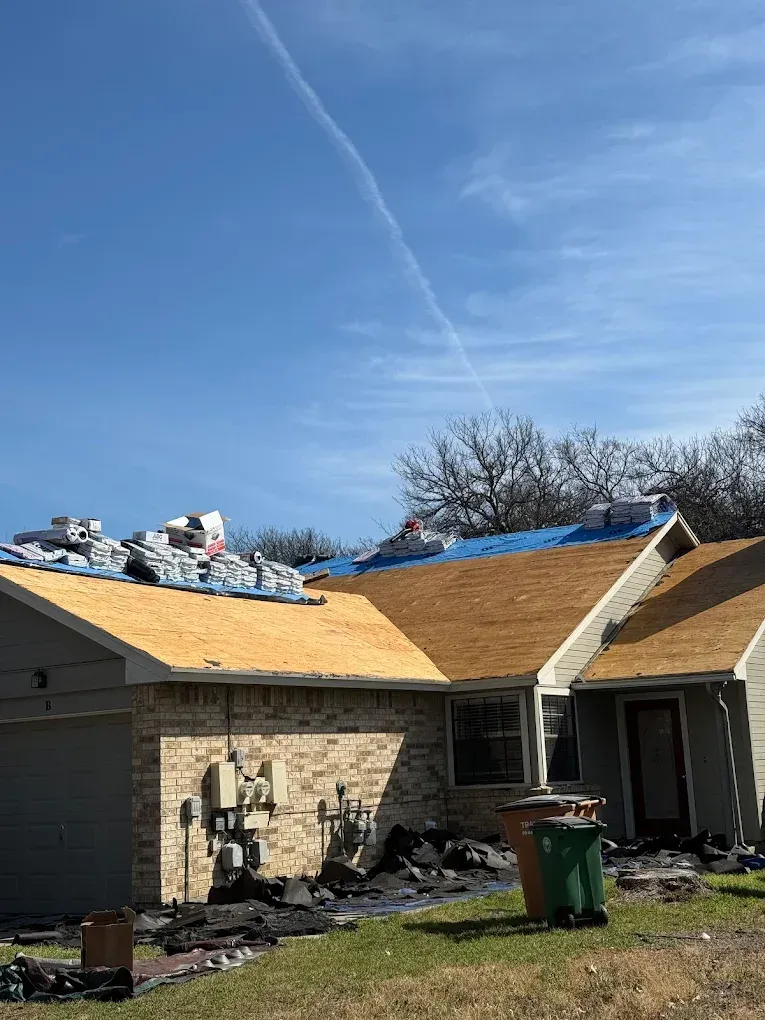 Partially roofed house under repair with plywood, debris in yard, and blue sky overhead