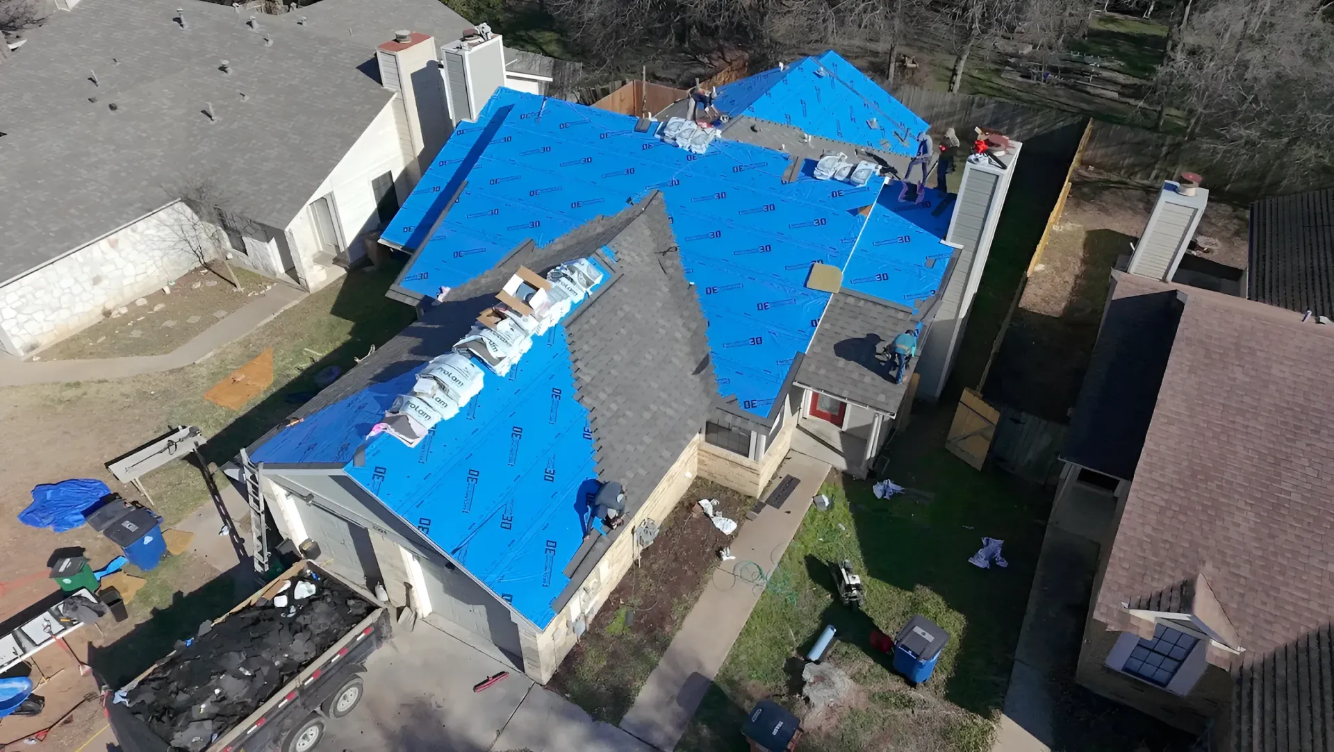 Aerial view of a building with a bright blue tarp covering most of its roof, surrounded by neighboring houses.