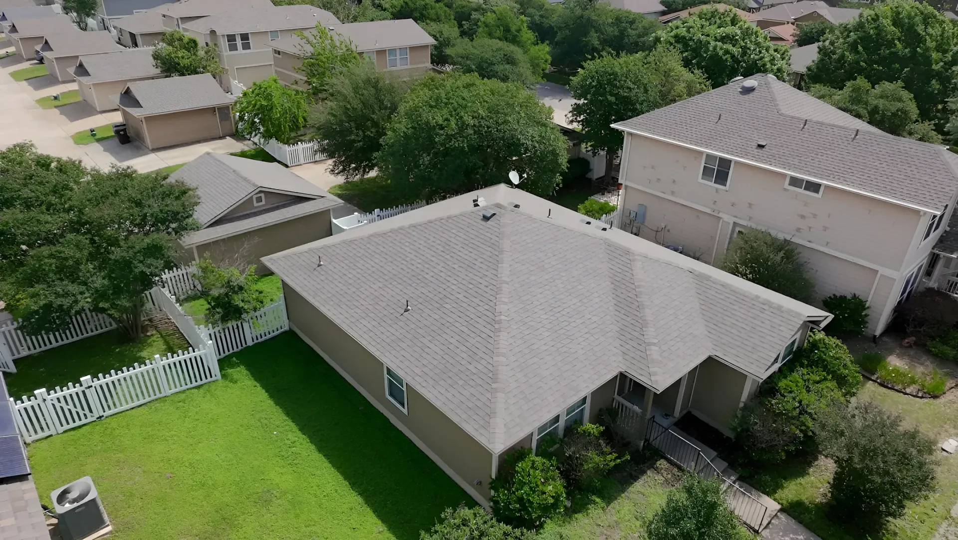 Aerial view of suburban houses with gray roofs, green lawns, and trees in a neighborhood.