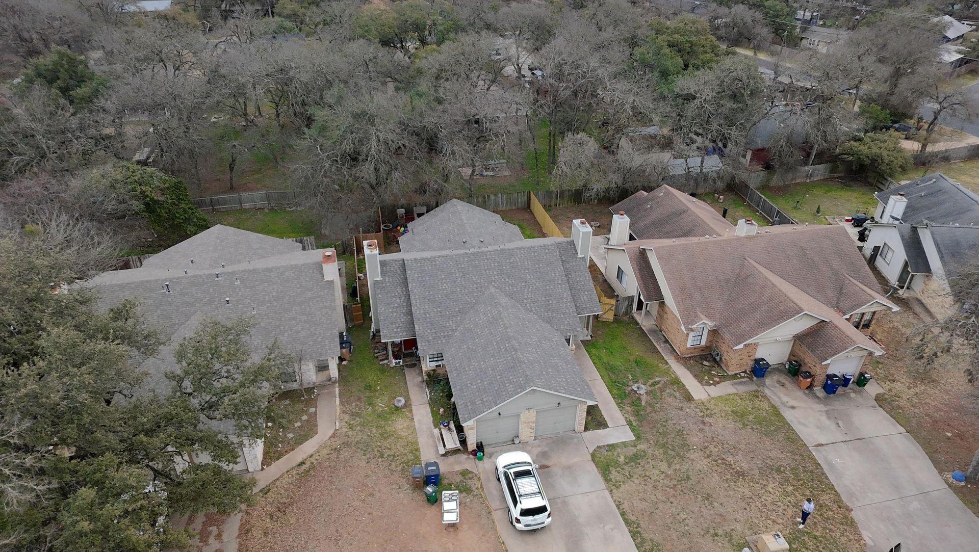 Aerial view of three houses with gray roofs beside trees and parked cars in a rural neighborhood