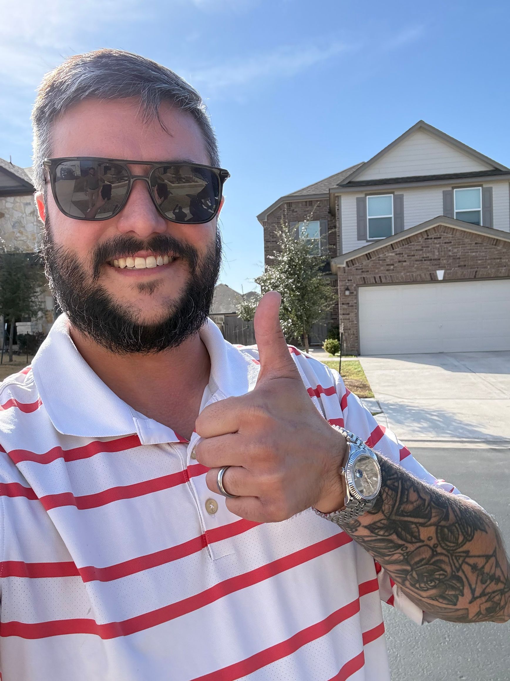 Man smiling outdoors in sunglasses, giving a thumbs-up in front of a suburban house.