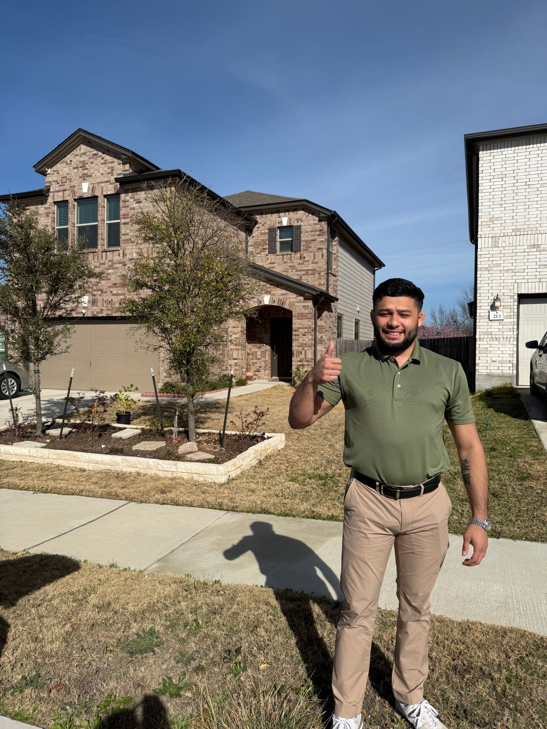 Man standing outside a suburban house, smiling and giving a thumbs-up on a sunny day.