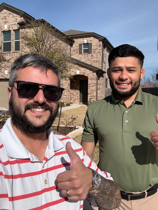 Two men smiling and giving thumbs up in front of a stone house on a sunny day
