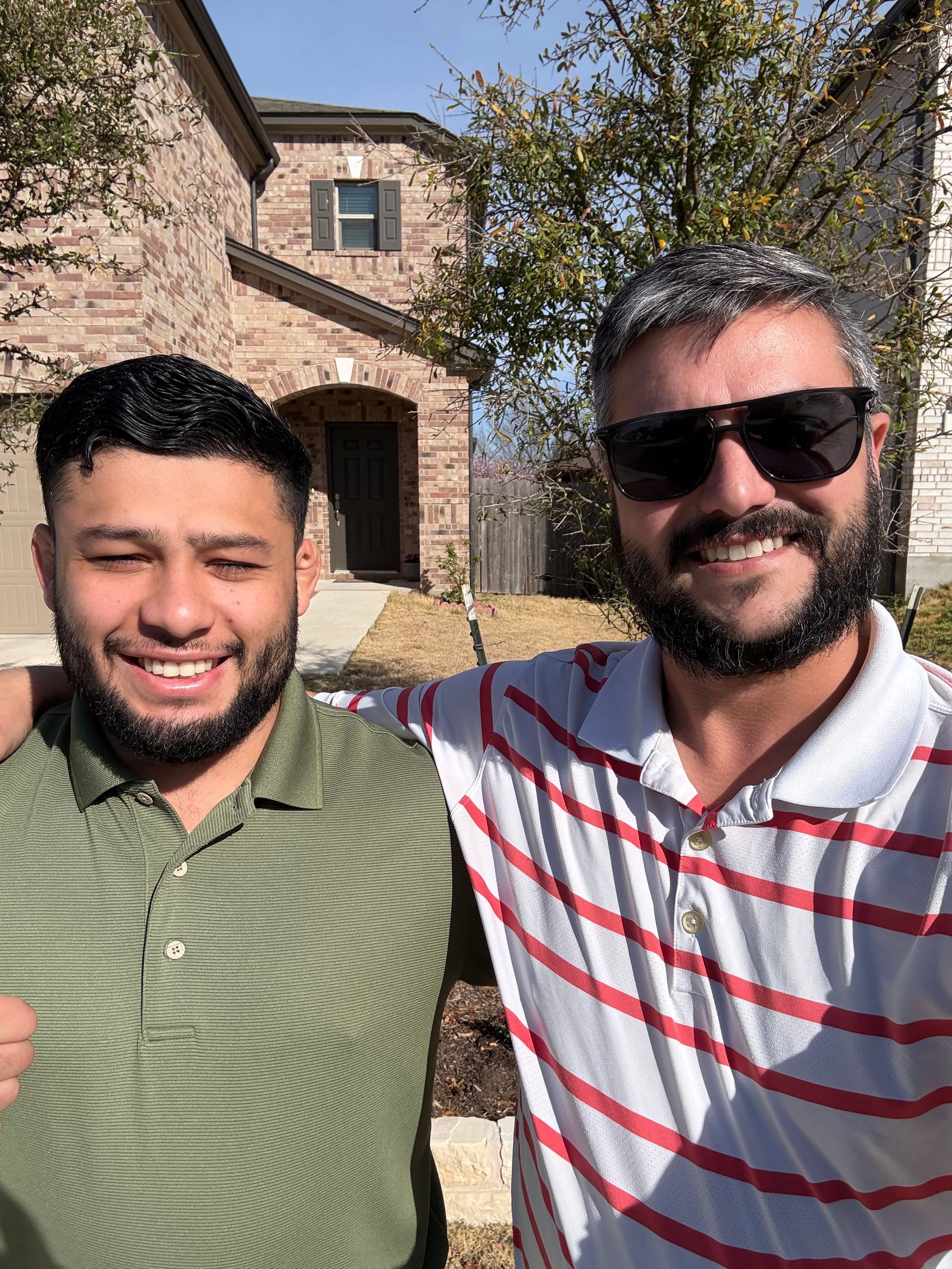 Two smiling people standing outdoors in front of a stone house in bright sunlight