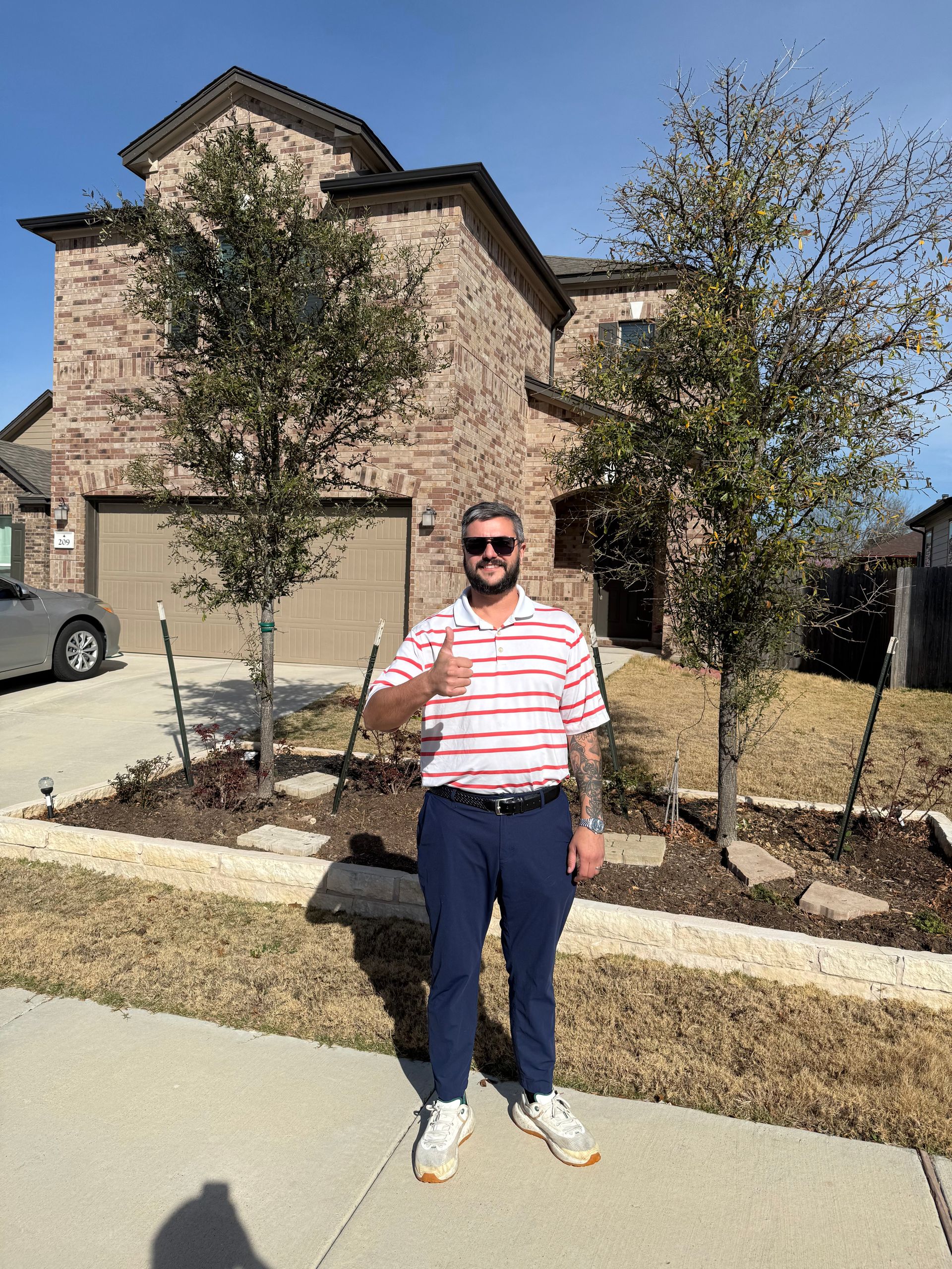 Man standing on a sidewalk in front of a brick house, holding a drink and wearing sunglasses.