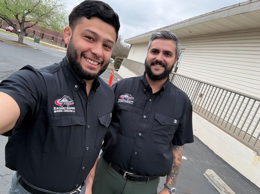 Two men in black shirts smiling for a selfie outdoors beside a building and railing.