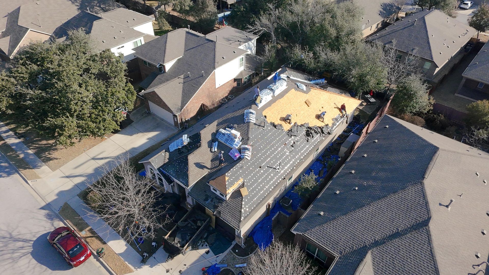 Aerial view of a house roof under repair, with workers and yellow tarp in a residential neighborhood.