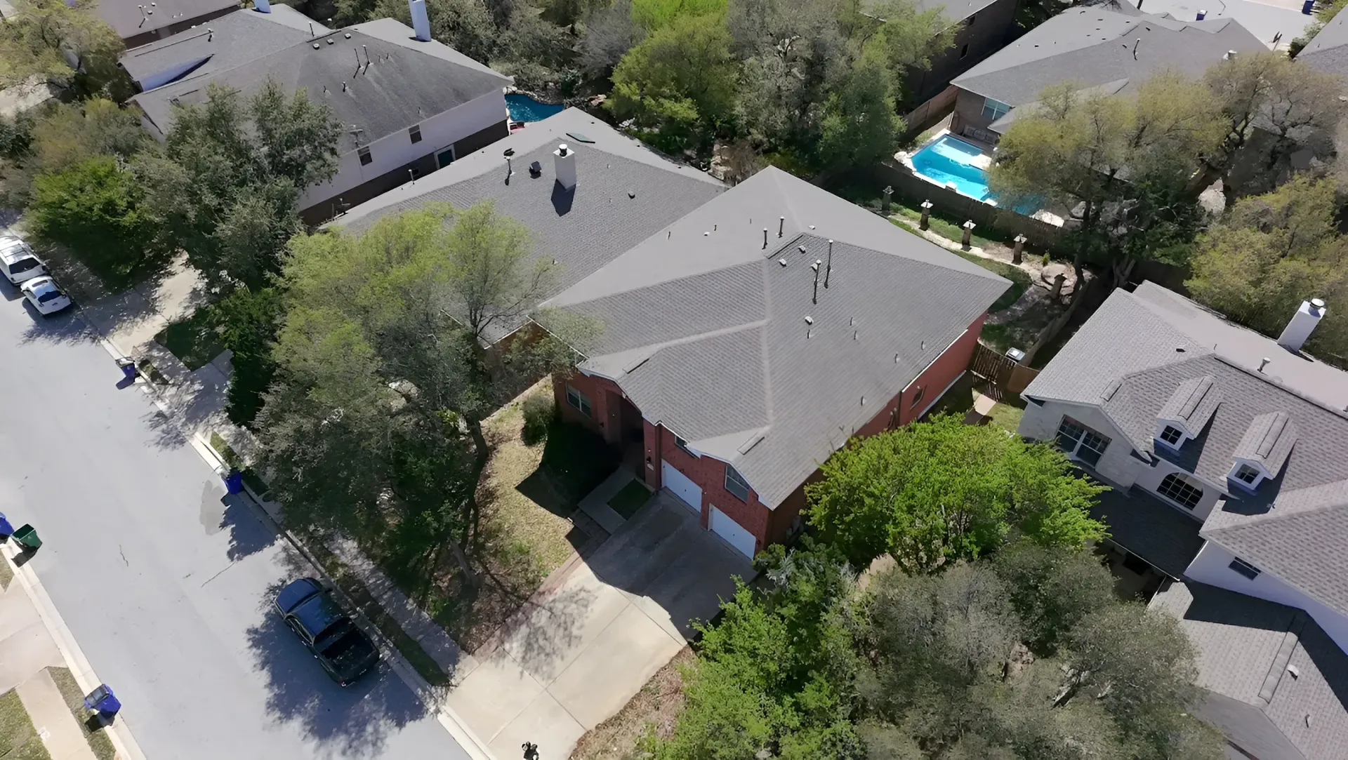 Aerial view of suburban homes with trees, parked cars, and a backyard swimming pool.